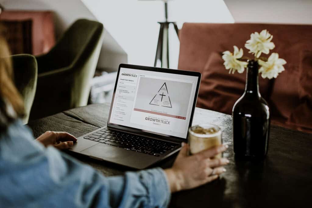 Person engaging with a laptop displaying the Growth Track program, featuring the Discipleship Triangle graphic, with a drink and a vase of flowers on a table in a cozy setting.