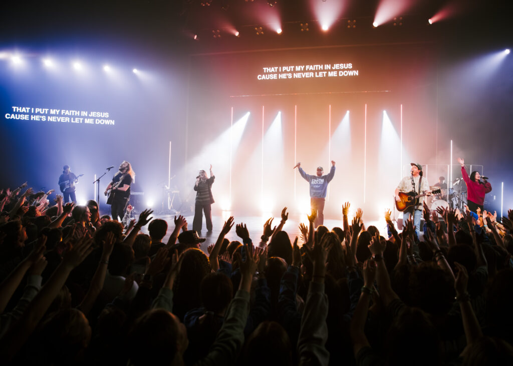 Worship band performing on stage at ONE WKND 2026, with audience raising hands, illuminated by stage lights and lyrics displayed, emphasizing the theme "INTO THE LIGHT."